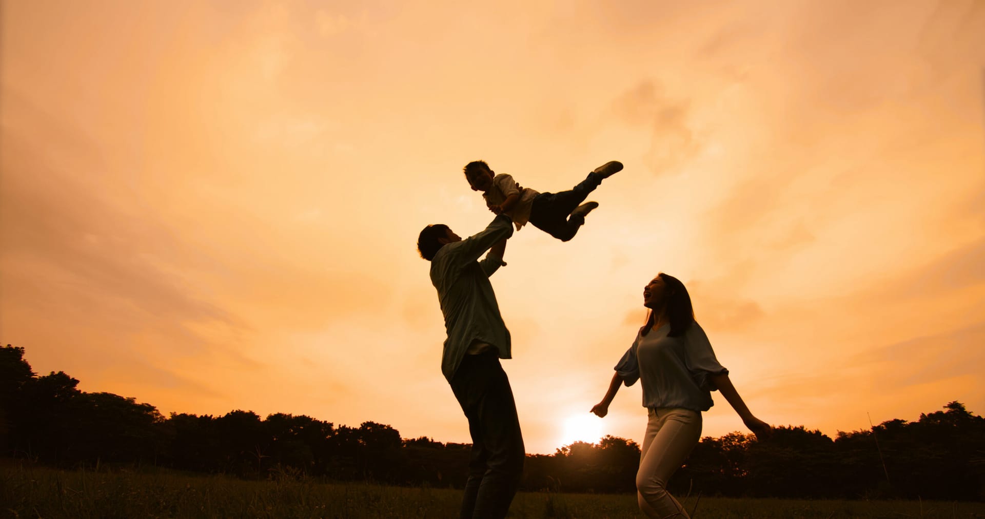 Asian parents holding child high in the air, sunset in the background.